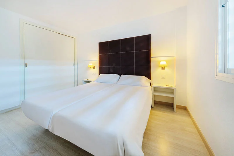 Habitaci&oacute;n moderna con cama doble blanca, mesita y pared oscura al fondo