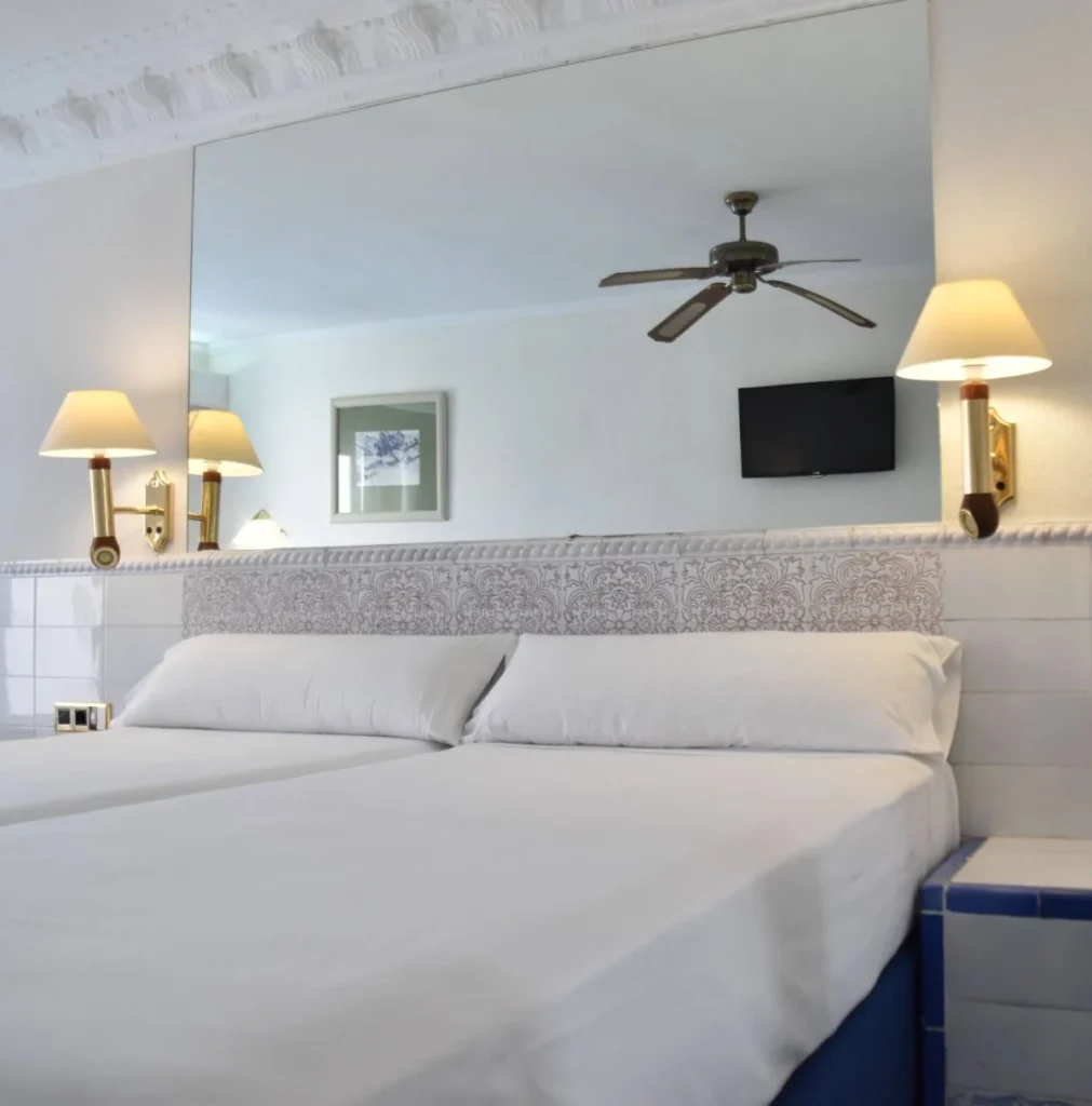 Habitaci&oacute;n de hotel con cama blanca, TV, ventilador y l&aacute;mparas de pared