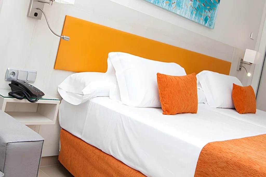 Cama blanca con almohadas naranjas en habitaci&oacute;n moderna.