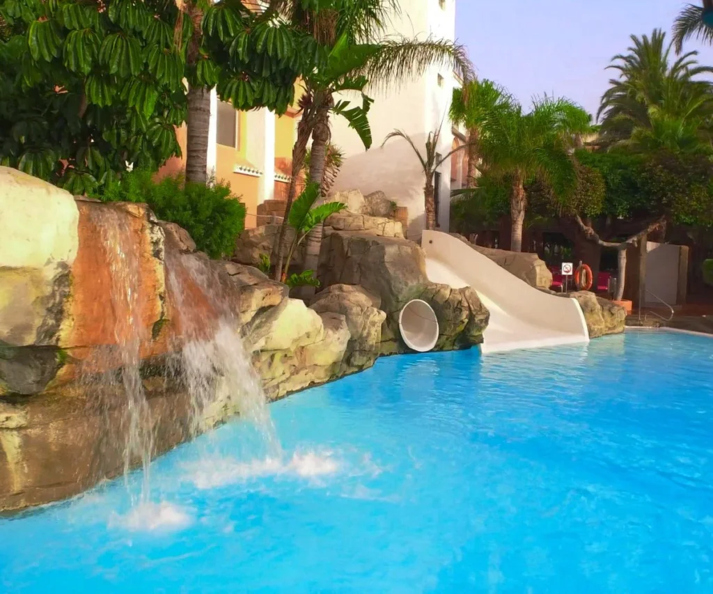 Piscina azul con cascada de piedra, palmeras y tobog&aacute;n blanco en hotel.