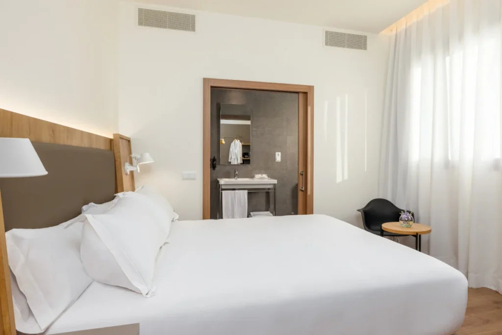 Habitaci&oacute;n moderna con cama blanca, mesita y espejo con ba&ntilde;o al fondo