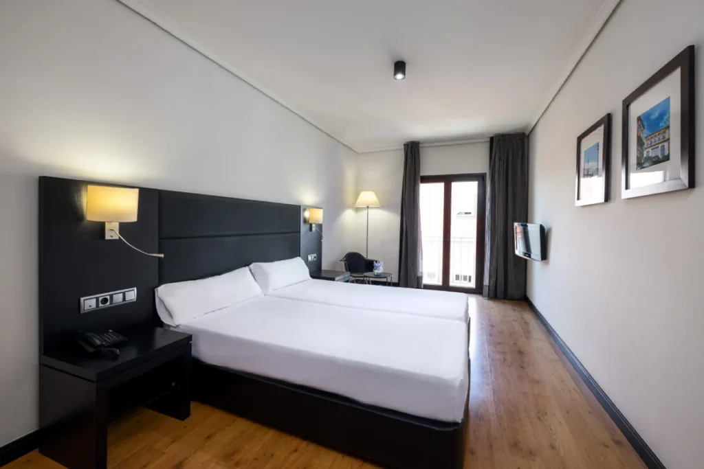 Dormitorio moderno con cama doble, mesitas y ventanas con cortinas oscuras.