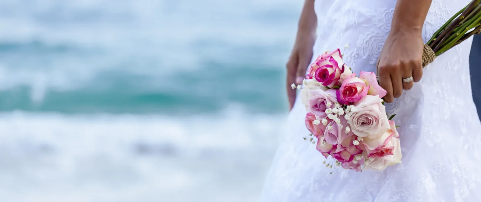 Novia sostiene ramo de rosas rosas y blancas junto al mar, con cielo azul