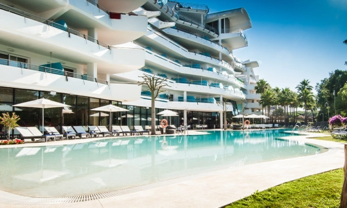 Edificio moderno junto a piscina rodeada de tumbonas y palmeras en un d&iacute;a soleado.