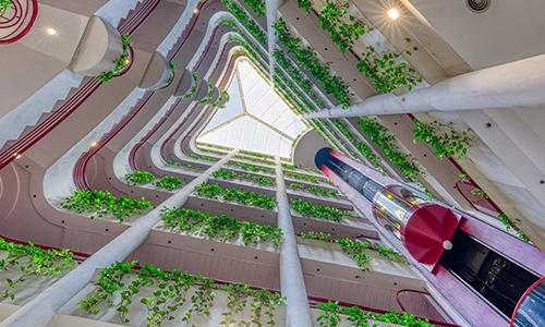 Vista desde abajo de un atrio con plantas y ascensores en un edificio moderno.