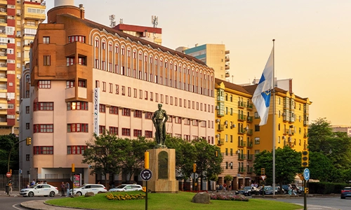 Edificio cl&aacute;sico y bandera en plaza con estatua al atardecer.