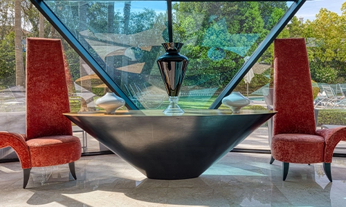 Mesa moderna con jarr&oacute;n y sillas rojas junto a ventana triangular.