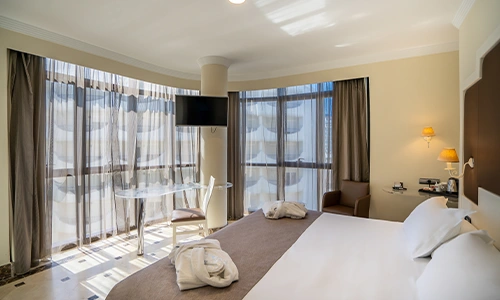 Habitaci&oacute;n de hotel luminosa con cama, televisor y ventanas grandes.
