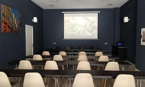 Aula vac&iacute;a con sillas, mesas y proyector encendido en la pared frontal.