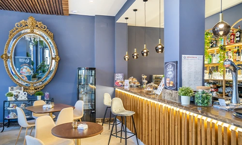 Cafeter&iacute;a moderna con mesas, espejo decorativo y barra iluminada.