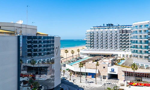 Vista de un hotel frente al mar con edificios y un estacionamiento cercano.