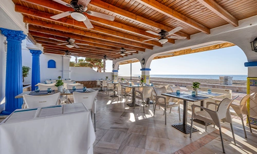 Terraza de restaurante junto al mar, con mesas y sillas bajo un techo de madera.
