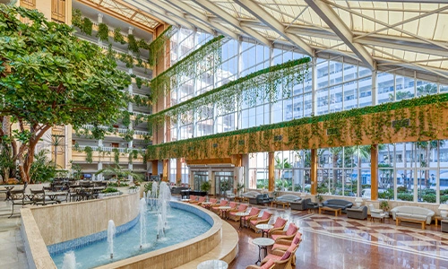 Lobby moderno con fuente, plantas y muebles elegantes en tonos c&aacute;lidos.