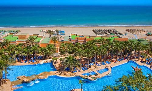 Vista de un complejo hotelero con piscinas y playa al fondo, cielo despejado.