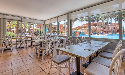 Restaurante luminoso con vistas a piscina y palmeras, sillas blancas y mesas dispuestas.
