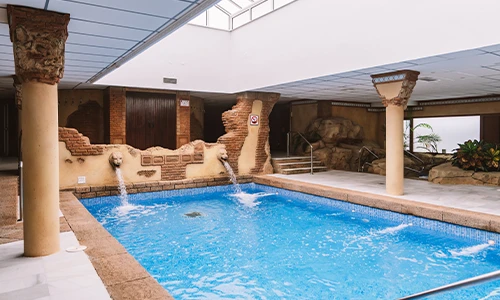 Piscina interior con columnas y cascadas decorativas en un ambiente r&uacute;stico.