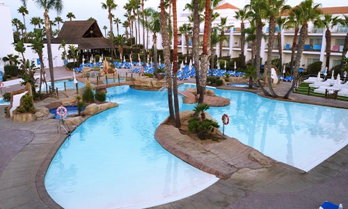 Piscina de hotel rodeada de palmeras y sillas azules bajo sombrillas blancas.