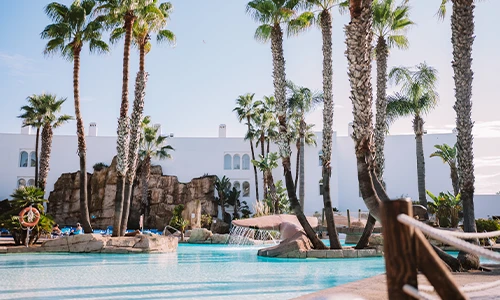 Piscina azul rodeada de palmeras y un edificio blanco al fondo.