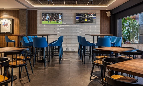 Bar deportivo con pantallas de TV, sillas azules y mesas de madera.