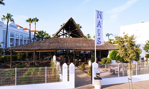 Edificio de restaurante con techo de paja y banner que dice "NAOS".