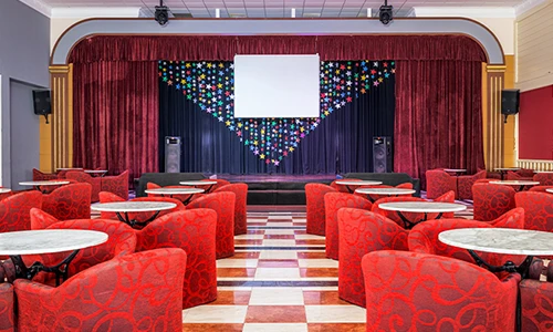 Sala con mesas y sillas rojas frente a un escenario con cortinas y pantalla.