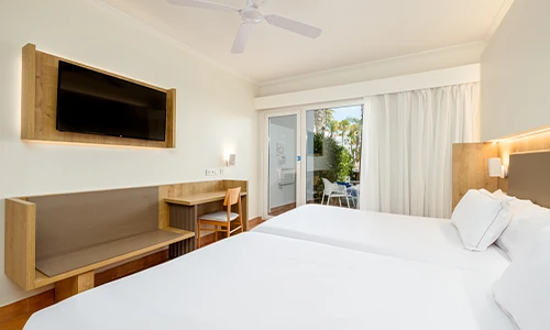 Habitaci&oacute;n de hotel moderna con dos camas y balc&oacute;n con vista al jard&iacute;n.