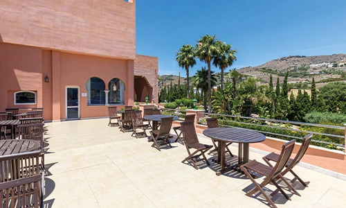Terraza con mesas y sillas, vista a palmeras y monta&ntilde;as bajo cielo azul.