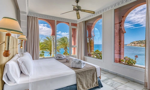 Habitaci&oacute;n luminosa con vista al mar y palmeras a trav&eacute;s de grandes ventanas.