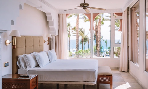 Habitaci&oacute;n luminosa con cama, vista al mar y palmeras a trav&eacute;s de las ventanas.