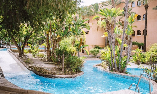 Piscina con vegetaci&oacute;n tropical y edificaciones en el fondo.