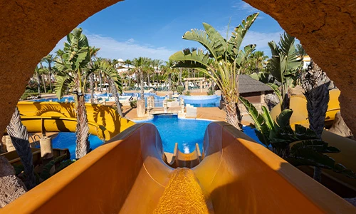 Tobog&aacute;n de agua amarillo en un parque con palmeras y piscinas al fondo.
