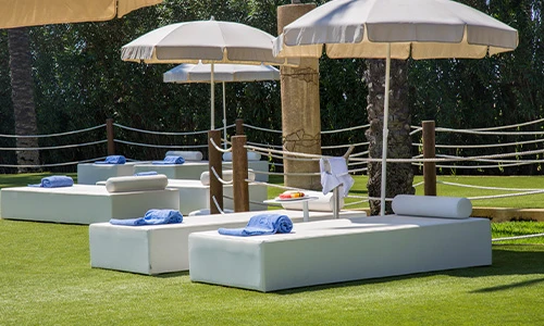 Camastros blancos con toallas azules bajo sombrillas en c&eacute;sped.