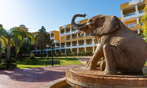 Estatua de elefante en fuente, con edificio amarillo a lo lejos y &aacute;rboles alrededor.