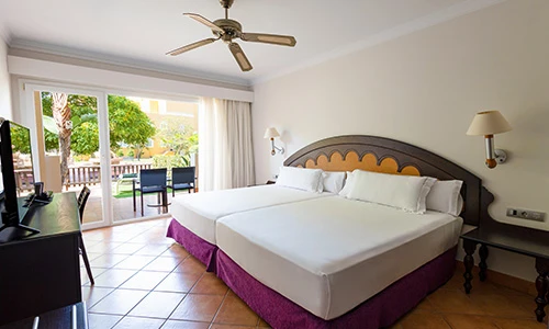 Habitaci&oacute;n de hotel con dos camas, ventilador y vista al jard&iacute;n.