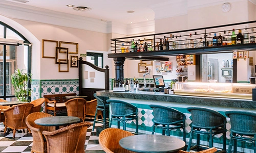 Bar acogedor con sillas de mimbre, barra y mesas; estilo cl&aacute;sico y luminoso.