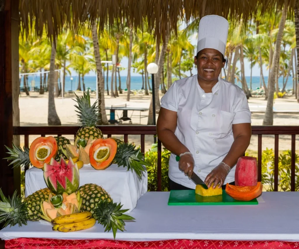 Chef con sombrero blanco corta fruta en mesa frente al mar y palmeras