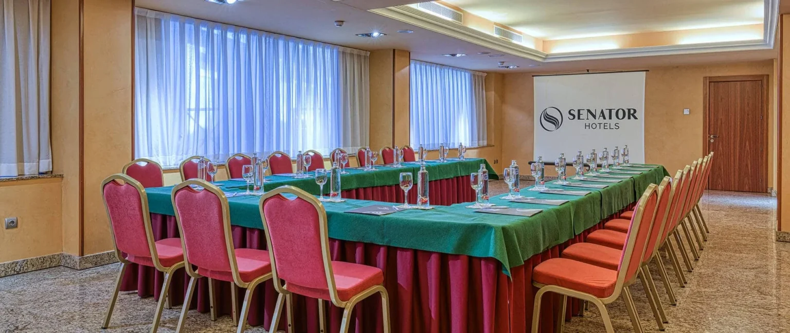 Sala de reuniones con mesas servidas, sillas rojas y pantalla Senator Hotels