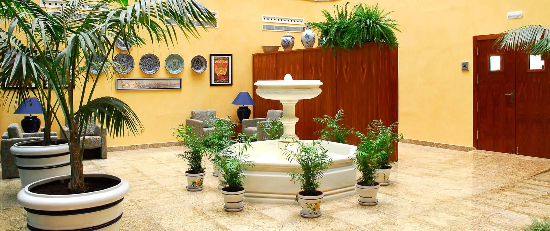 Patio interior con fuente blanca, plantas y puertas de madera color marr&oacute;n.