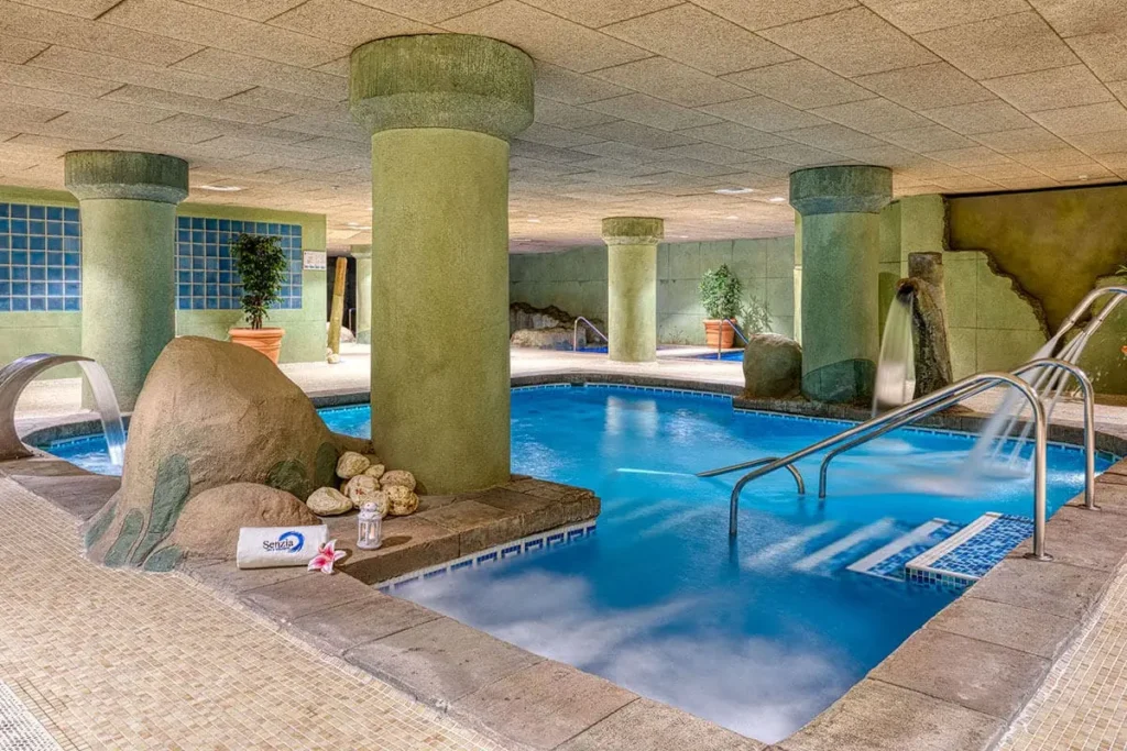 Piscina interior con chorros, columnas verdes y &aacute;rea de descanso con tumbonas