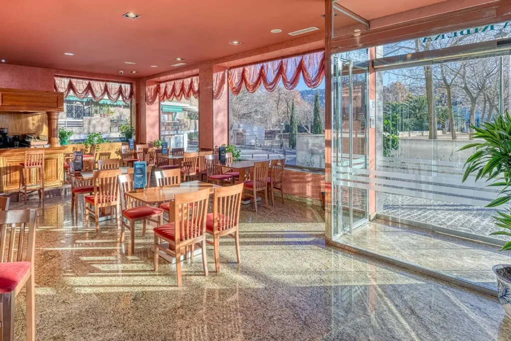 Sal&oacute;n con mesas y sillas de madera, cortinas rojas y ventanales con jard&iacute;n.
