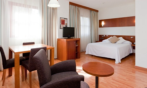 Habitaci&oacute;n hotel moderna con cama, mesa, sillas, TV y cortinas luminosas.