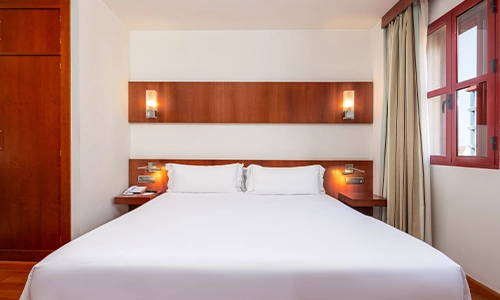 Habitaci&oacute;n de hotel con cama doble, luz c&aacute;lida y ventana con cortinas.