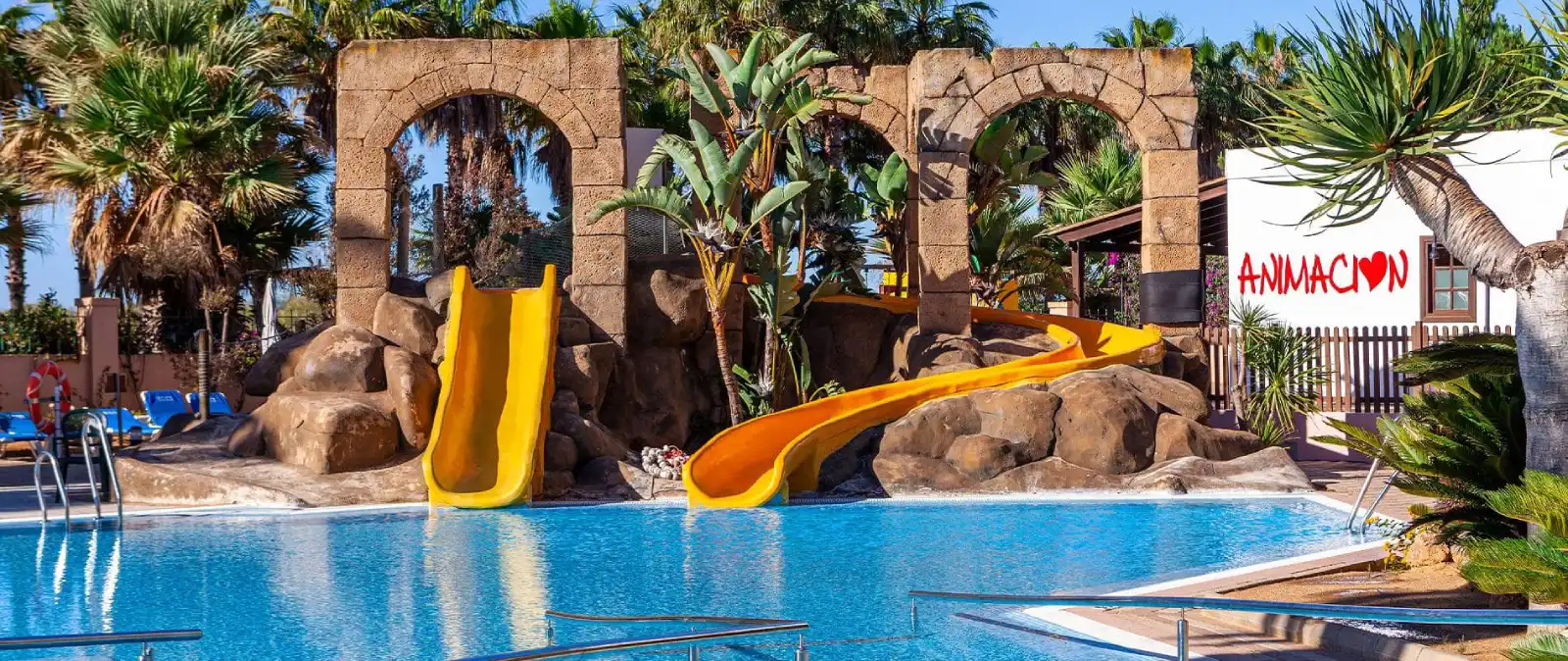 Piscina de hotel con toboganes amarillos y arco de piedra entre palmeras.