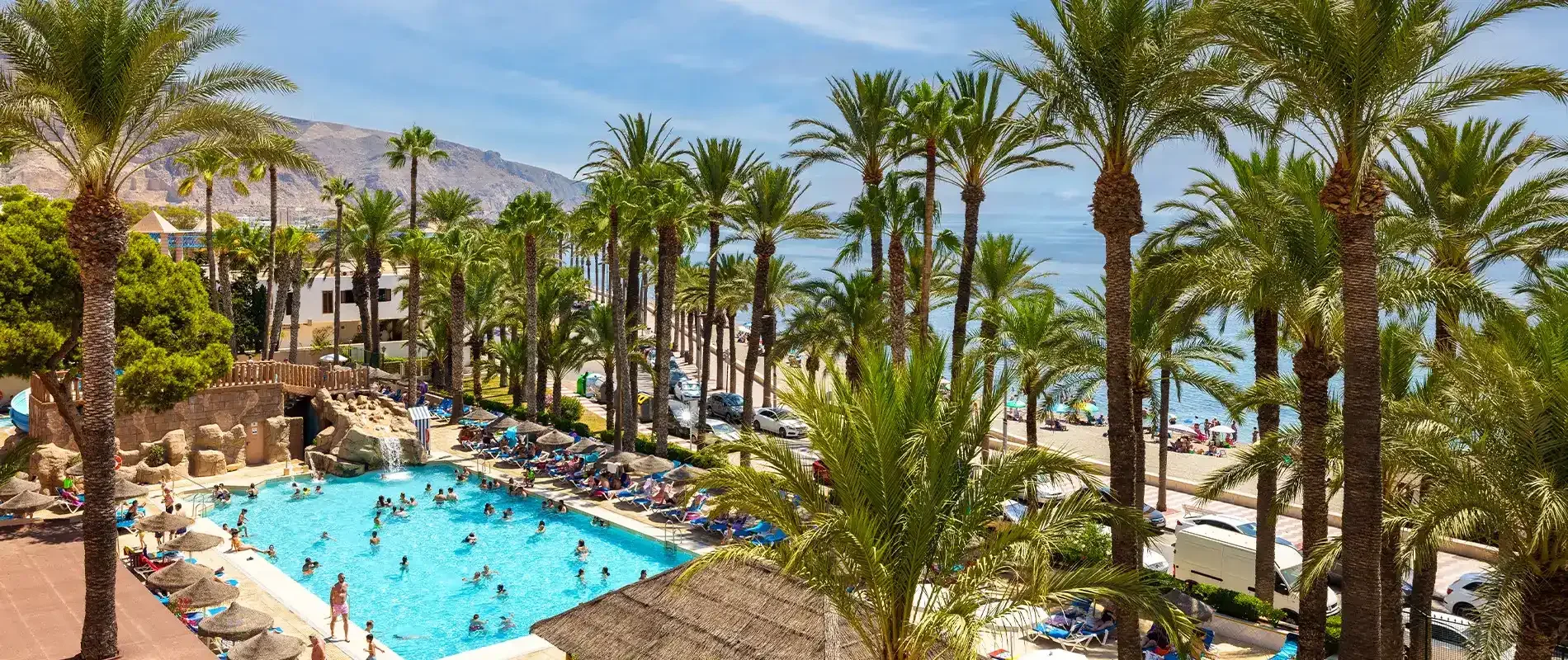 Piscina junto al mar con palmeras y monta&ntilde;as al fondo, resort vacacional