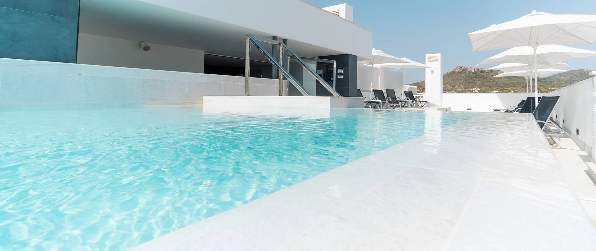 Piscina moderna con tumbonas y sombrillas blancas, junto a edificio blanco.