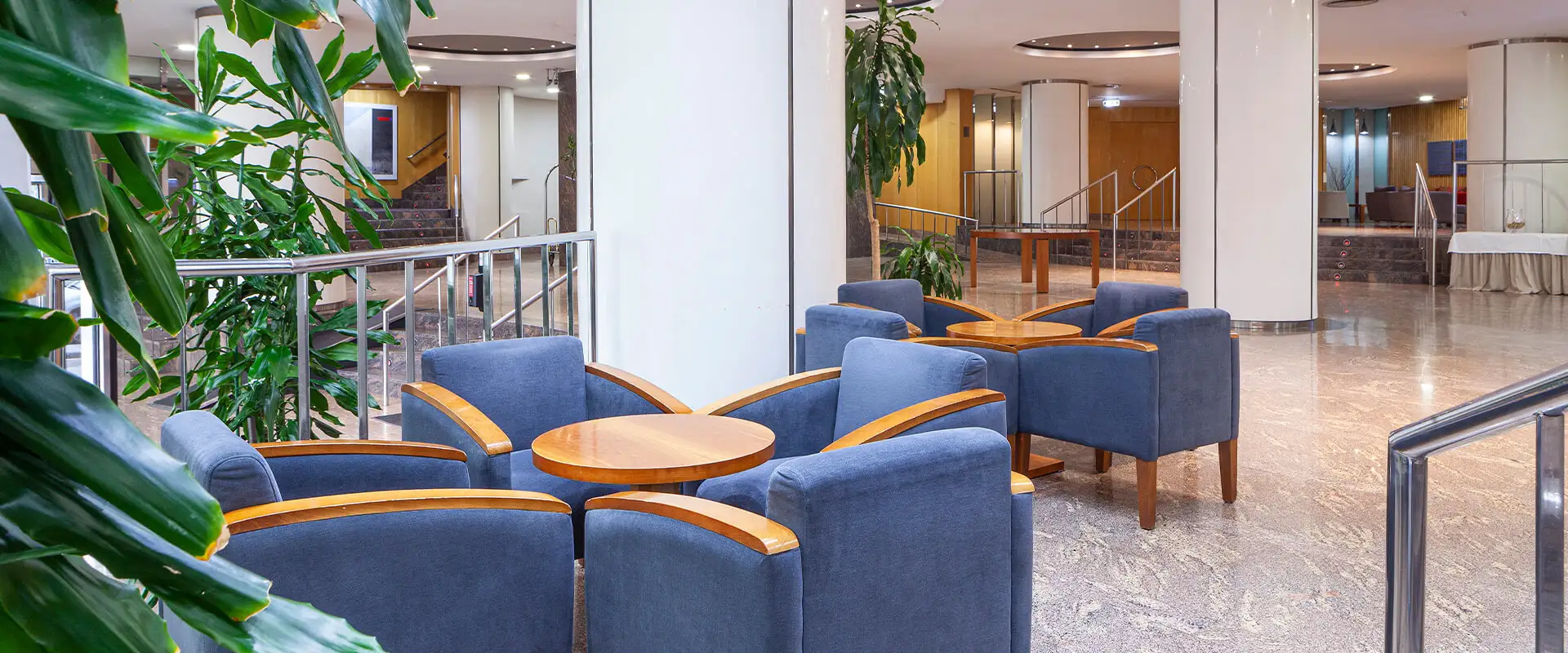 Sala de recepci&oacute;n con sof&aacute;s azules, mesas de madera y plantas en un vest&iacute;bulo interior