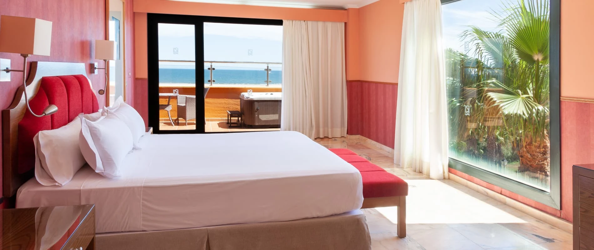 Habitaci&oacute;n de hotel con cama roja, cortinas y vista al mar y jard&iacute;n tropical