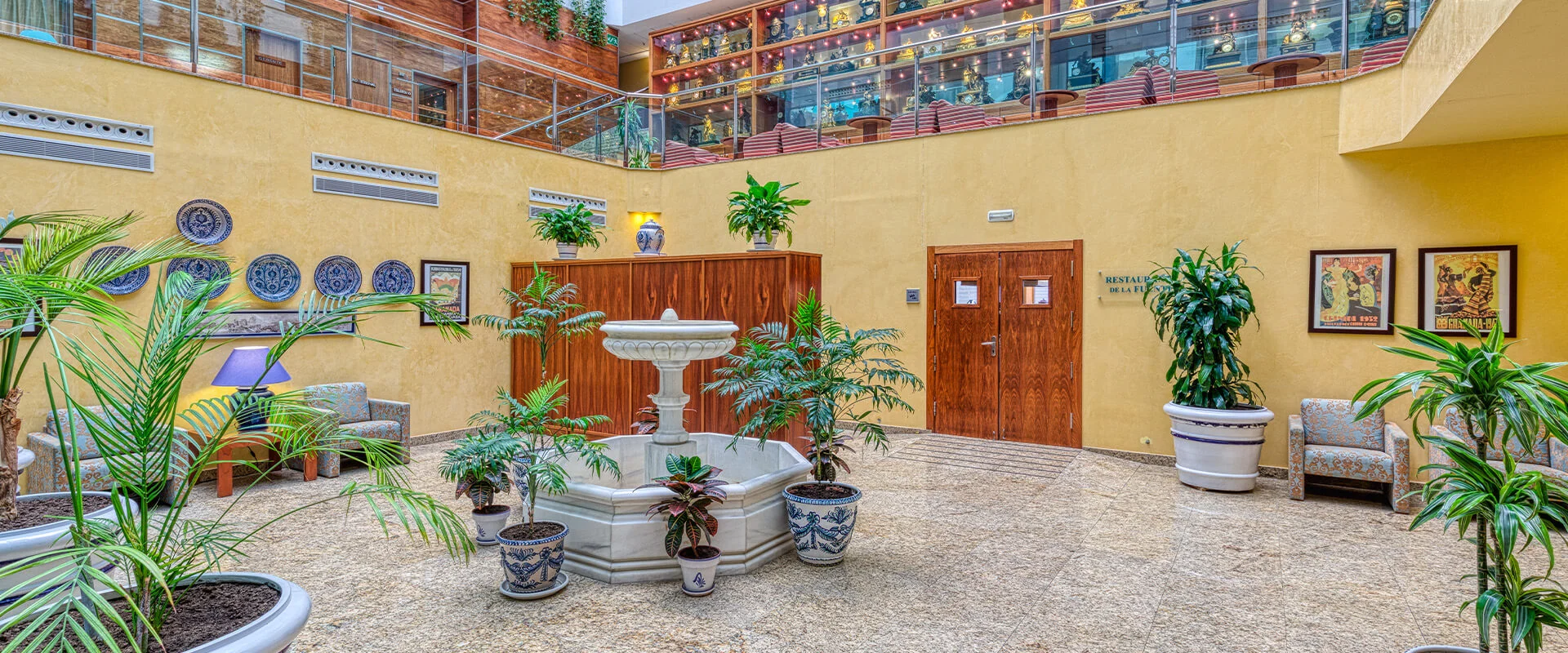 Patio interior con fuentes, plantas y sillones, rodeado por balc&oacute;n con luces