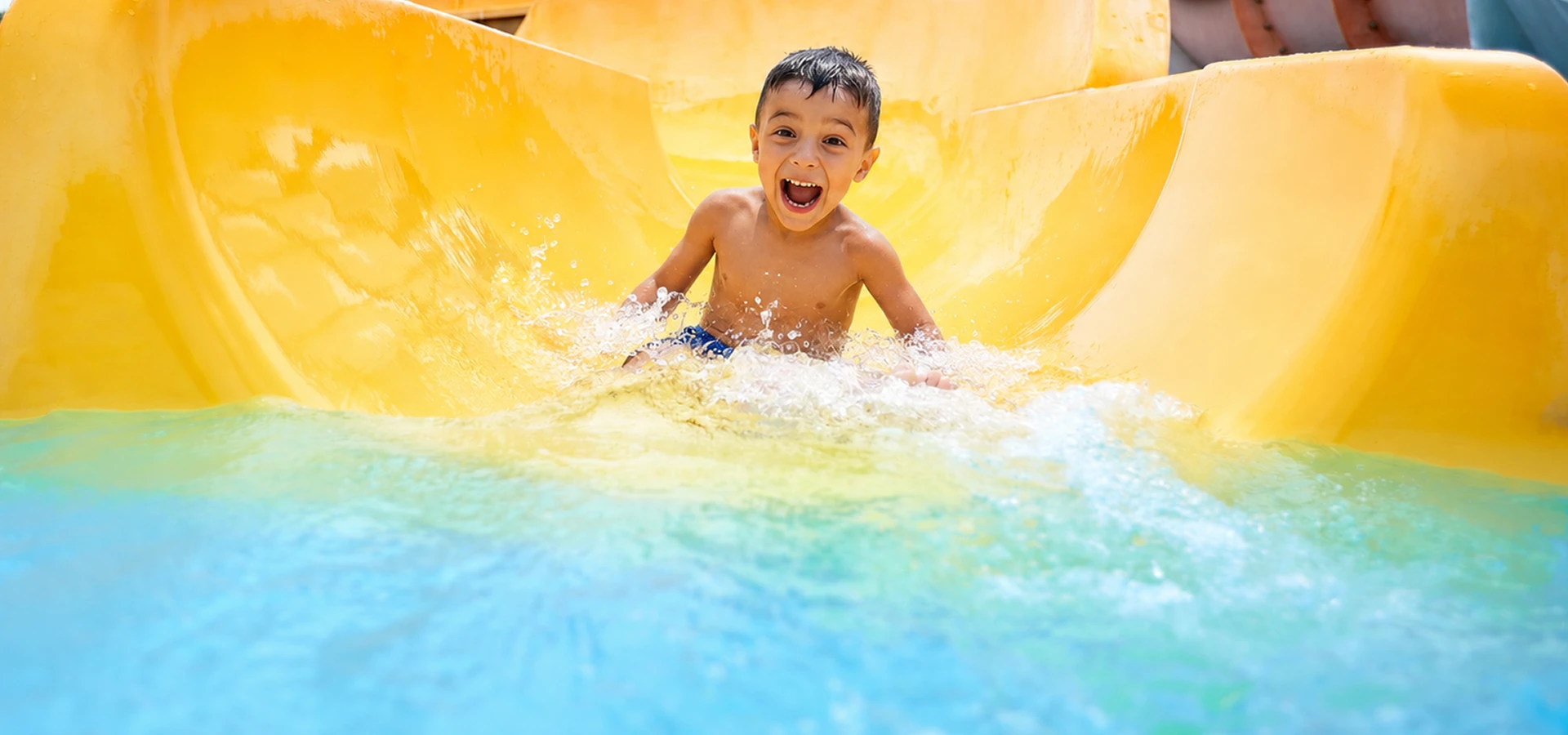 Ni&ntilde;o sonriente jugando en el tobog&aacute;n amarillo con salpicaduras de agua