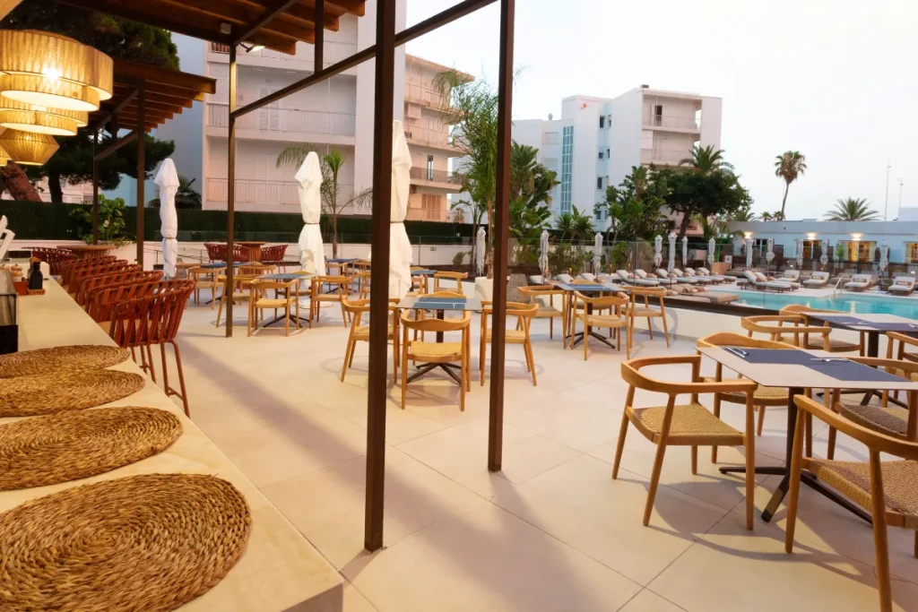 Terraza de hotel con mesas, sillas de madera y piscina al fondo.